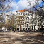 Sonniger Stadtplatz mit kahlen Bäumen, umgeben von Gebäuden und geparkten Autos. Fußgänger und Sitzgelegenheiten im Freien sind sichtbar.