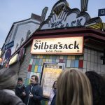 Eine Gruppe von Menschen vor der Kneipe „Zum Silbersack“ mit Neonlichtern, bunten Fliesen und Straßenschildern im Hintergrund.