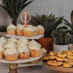 Cupcakes mit weißem Zuckerguss und Chocolate Chip Cookies auf Ständern, umgeben von Topfpflanzen, bilden die perfekte Kulisse für eine stimmungsvolle Junggesellenabschiedsfeier.