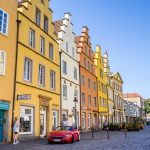 Farbenfrohe historische Gebäude säumen eine Kopfsteinpflasterstraße, davor parkt unter einem klaren blauen Himmel ein rotes Auto.