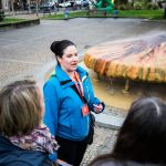 Ein Reiseleiter spricht zu einer kleinen Gruppe vor einem einzigartigen, pilzförmigen Brunnen in einer städtischen Umgebung.