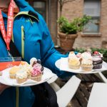 Person, die Teller mit verschiedenen Cupcakes hält, eine blaue Jacke und ein rotes Schlüsselband im Freien trägt.