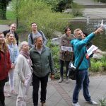 Eine Gruppe von Menschen steht im Freien, eine Person zeigt auf einen Teich mit Springbrunnen.