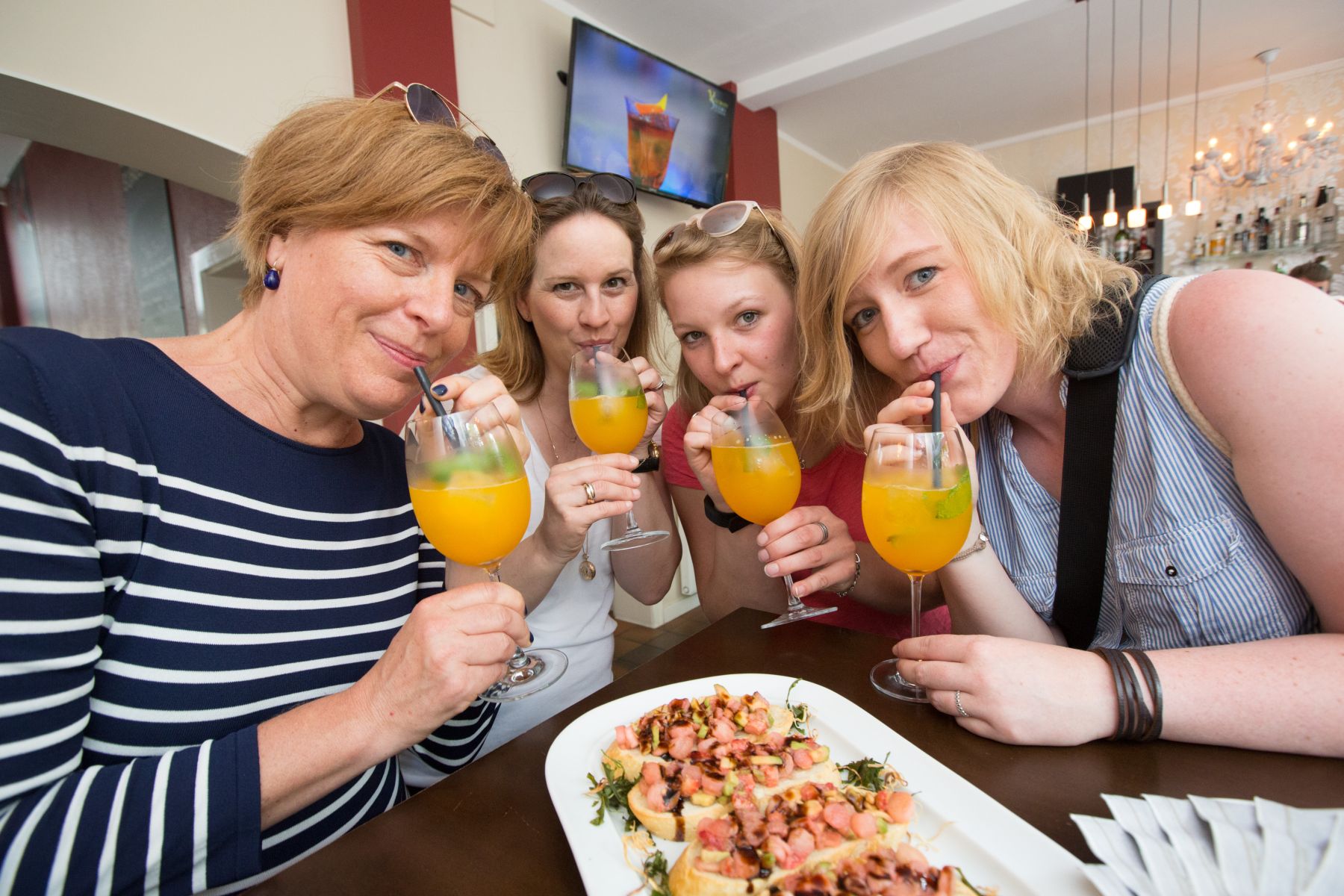 Vier Frauen erfreuen sich an einer wunderbaren Geschenkidee, genießen Orangengetränke und Häppchen an einer Bar und lächeln herzlich in die Kamera.