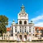 Großartiges historisches Gebäude mit reich verzierter Fassade, drei Kuppeltürmen und einem großen Baum vor einem klaren blauen Himmel.