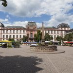 Ein großes historisches Gebäude mit einem Platz, einem Brunnen und Marktständen unter einem bewölkten Himmel.