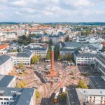 Die Luftaufnahme von Darmstadt zeigt eine geschäftige Stadt mit einem zentralen Platz, umliegenden Gebäuden und einer Eisenbahnlinie, die sich unter einem teilweise bewölkten Himmel hindurchschlängelt.