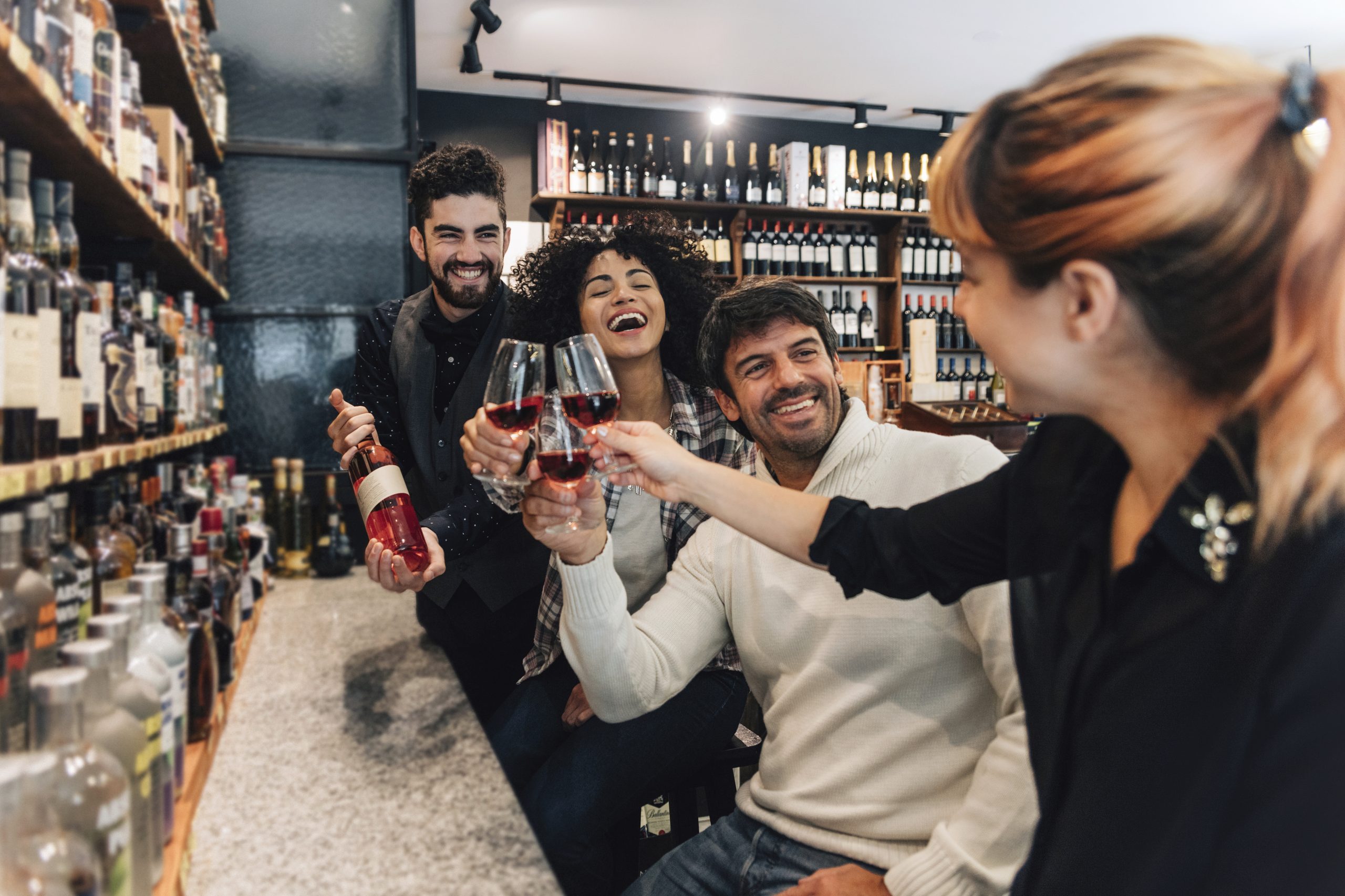 Vier Freunde stoßen mit Weingläsern an einer Bar an, umgeben von Regalen voller Weinflaschen. Sie lächeln und haben Spaß und fangen die festliche Stimmung einer Weihnachtsfeier in vollem Gange ein.