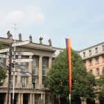 Ein historisches Gebäude mit Statuen darauf, umgeben von Bäumen und einem Wegweiser mit mehreren Richtungsangaben zur Stadt.