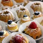 Mini-Gugelhupfe mit Beeren und Puderzucker, serviert in Papierförmchen auf einem Tablett.