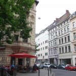 Straßenansicht mit Straßencafé, Menschen sitzen unter einem roten Sonnenschirm, umgeben von historischen Gebäuden und geparkten Autos.