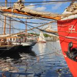 Segelboote und ein rotes Schiff mit Anker liegen in einem sonnigen Hafen.