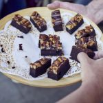 Ein Holztablett mit Schokoladenbrownies, garniert mit Nüssen auf einem Spitzendeckchen. Eine Hand greift nach einem Stück.