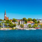 Der Küstencharme Flensburgs erstrahlt mit Segelbooten im Hafen, farbenfrohen Gebäuden und einer Kirche mit einem markanten Turm unter einem klaren blauen Himmel.