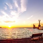 Sonnenuntergang über einem See mit Bänken, einem Pier und einem vor Anker liegenden Schiff unter einem leuchtenden Himmel.