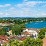 Luftaufnahme der malerischen Seestadt Konstanz mit ihren rot gedeckten Gebäuden, üppigem Grün und einem klaren blauen See, der unter einem sonnigen Himmel glitzert.