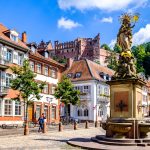 Historischer europäischer Platz mit einem Brunnen, farbenfrohen Gebäuden und einem Schloss auf einem Hügel unter einem blauen Himmel.