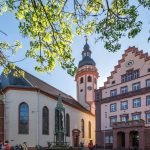 Historisches Gebäude mit detaillierter Architektur, einer Uhr und einem zentralen Turm, umgeben von Bäumen unter einem klaren Himmel.