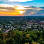 Luftaufnahme einer Stadt bei Sonnenuntergang mit bunten Wolken und einer Mischung aus städtischen Gebäuden und grünen Bäumen.