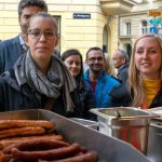 Menschen, die vor einem Imbissstand Schlange stehen, im Vordergrund sind Würstchen und Gewürze zu sehen.