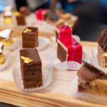 Verschiedene Gebäckstücke und Kuchen mit Schokoladen-, Obst- und Sahneüberzug auf einem Holztablett.