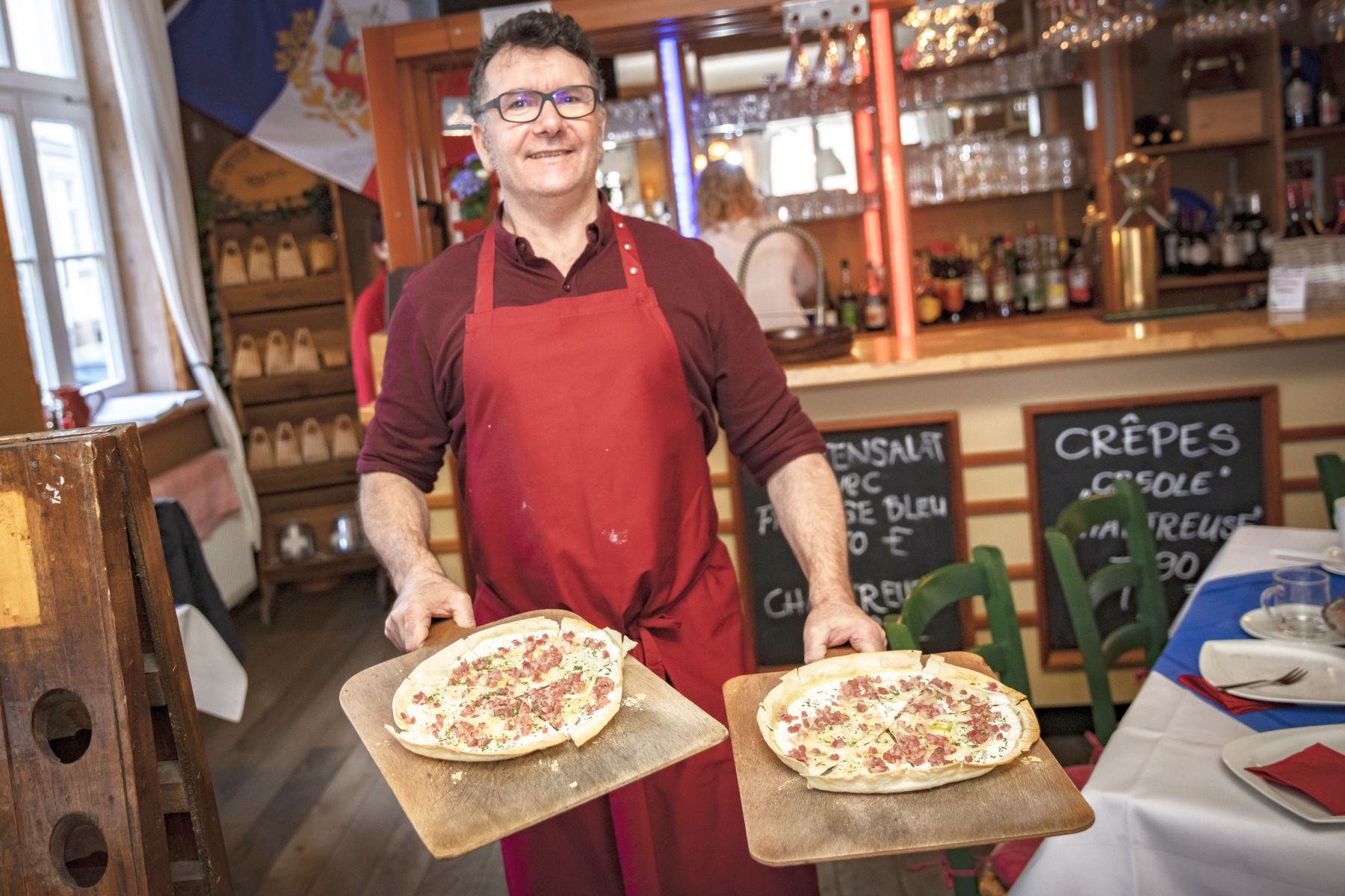 Lächelnder Mann in roter Schürze hält zwei Pizzen auf Holzbrettern in einem gemütlichen Restaurant, perfekt für Ihre Weihnachtsfeier.