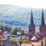 Malerische Aussicht auf eine Stadt mit Kirchtürmen aus rotem Backstein und einem üppig grünen Hügel im Hintergrund.