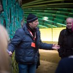 Ein Mann in einer blauen Jacke und einer Beanie-Mütze gestikuliert, während er zu einer kleinen Gruppe in einem mit Graffiti besprühten Tunnel spricht.