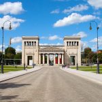 Neoklassizistisches Gebäude mit Säulen und Skulpturen, unter einem blauen Himmel mit Wolken, umgeben von grünen Rasenflächen und Wegen.