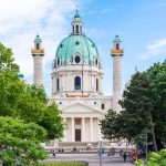 Die Karlskirche in Wien, umgeben von Bäumen, mit Menschen im Vordergrund unter einem klaren blauen Himmel.