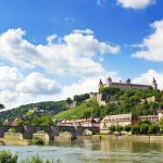 Malerische Aussicht auf die Festung Marienberg auf einem Hügel mit einer alten Steinbrücke über einen Fluss unter einem blauen Himmel mit Wolken.