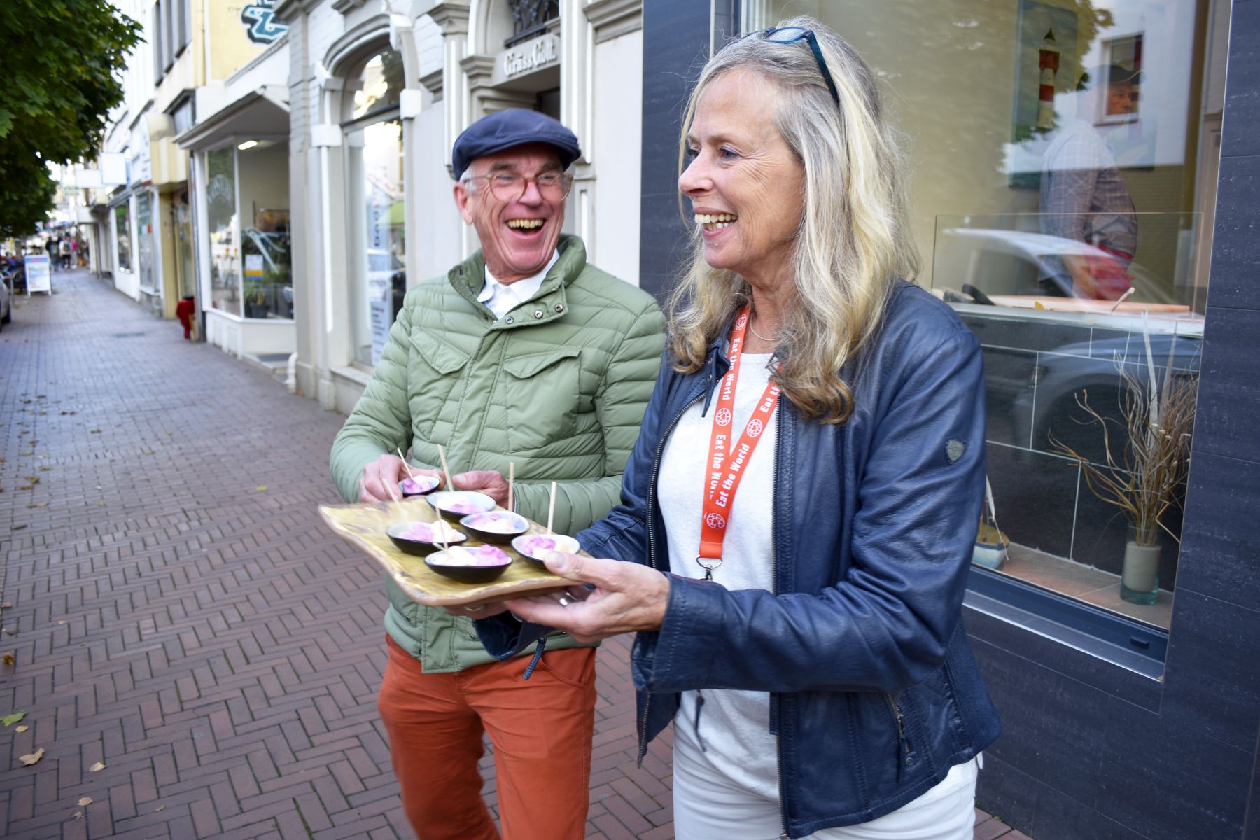 Zwei Menschen lachen und halten ein Holztablett mit kleinen Tassen auf einer Stadtstraße, aufgenommen während eines aufregenden Betriebsausflugs, und genießen gemeinsam die lebhafte Atmosphäre.