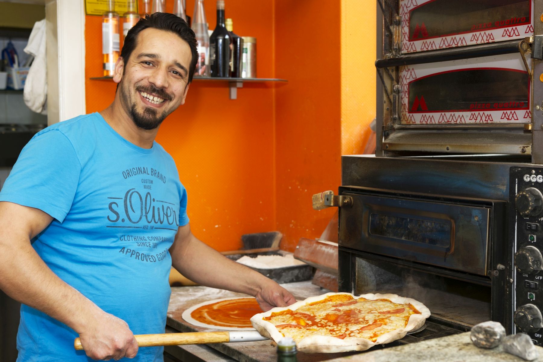 Während eines lebhaften Betriebsausflugs legt ein lächelnder Mann in einem blauen Hemd eine Pizza in einen Pizzaofen in der Küche.