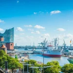 Panoramablick auf eine Hafenstadtlandschaft mit historischen Schiffen, modernen Gebäuden und blauem Himmel mit flauschigen Wolken.