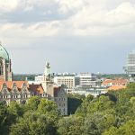 Skyline einer Stadt mit einem historischen Gebäude mit grüner Kuppel, modernen Wolkenkratzern und üppigen Bäumen im Vordergrund.