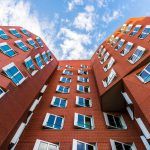 Stilisiertes rotes Backsteingebäude mit gekippten Fenstern vor einem teilweise bewölkten Himmel.