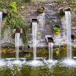 Eine Steinmauer mit kaskadierenden Wasserfontänen und Grünpflanzen in Metallgefäßen.