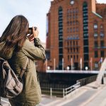 Frau mit Rucksack fotografiert an einem sonnigen Tag ein großes Backsteingebäude.