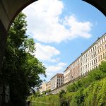 Blick unter einer Brücke auf einen von Gebäuden und Grün gesäumten Kanal unter einem blauen Himmel.