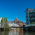 Ein malerischer Blick auf eine Metallbrücke über einen ruhigen Fluss, flankiert von modernen Gebäuden und üppigen Bäumen unter einem klaren blauen Himmel.