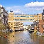 Menschen fahren Kajak auf einem Kanal, der von historischen Gebäuden und einer Skybridge umgeben ist, unter einem teilweise bewölkten Himmel.