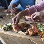 Menschen greifen nach verschiedenen Pizzen auf einem Holztisch und genießen die herrlichen kulinarischen Erlebnisse, neben ihnen stehen Weingläser.