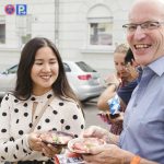 Zwei Menschen lächeln und halten kleine Teller mit Essen im Freien, im Hintergrund sind ein geparktes Auto und ein Gebäude zu sehen.