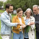 Vier Erwachsene stehen zusammen auf der Wiese im Berliner Stadtteil Friedrichshain und studieren konzentriert eine Karte. Sie sind leger in Jacken gekleidet und scheinen sich auf eine spannende Krimi-Rätseltour vorzubereiten. Üppige Bäume bilden eine malerische Kulisse.