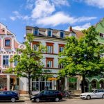 Unter einem strahlend blauen Himmel säumen farbenfrohe historische Gebäude eine Straße in einer europäischen Stadt, davor parken Autos.