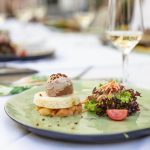 Gourmetgericht mit Pastete, Toast, Salat und einem Glas Weißwein auf einem grünen Teller an einem Restauranttisch.