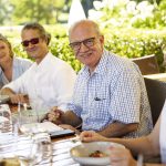 Vier Personen genießen eine Mahlzeit im Freien an einem Tisch, mit üppigem Grün im Hintergrund.