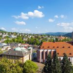 Malerische Aussicht auf eine europäische Stadt mit einer Kirche, üppigem Grün und Hügeln in der Ferne unter einem blauen Himmel.