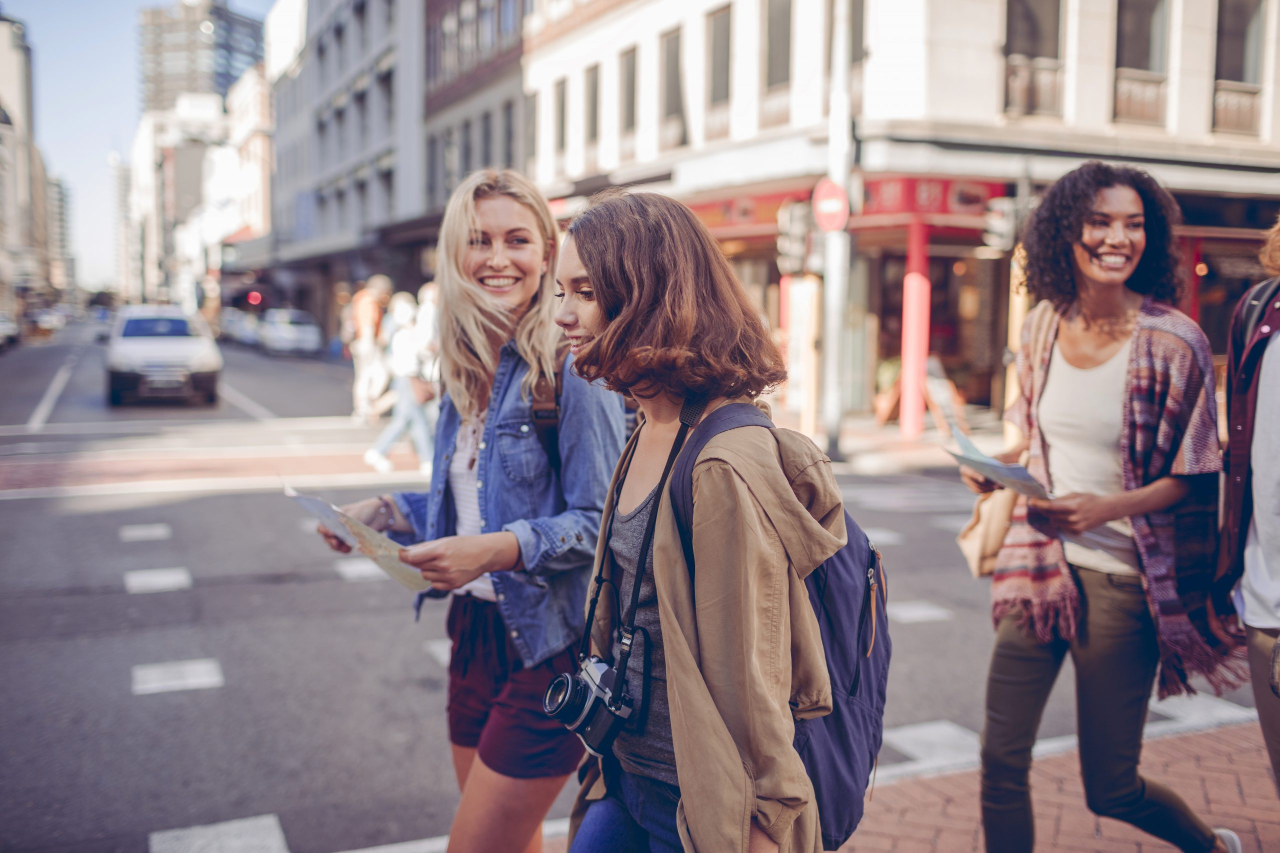 Vier Freunde überqueren auf einem Junggesellenabschied lächelnd und mit Stadtplänen in der Hand eine belebte Stadtstraße, umgeben von hoch aufragenden Gebäuden und Autos.