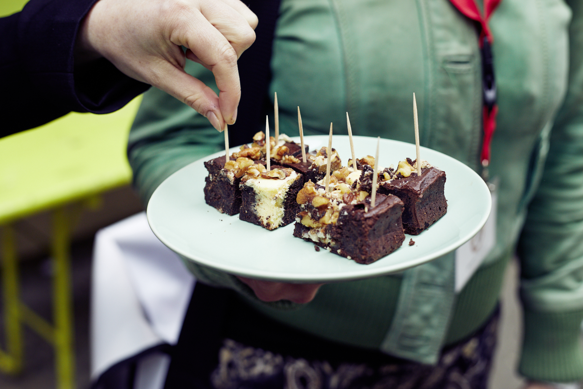 Beim lebhaften Betriebsausflug hält jemand einen Teller mit Schokoladenbrownies mit Nüssen und Zahnstochern in der Hand, während ein anderer gierig nach einem Stück greift.